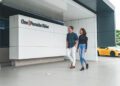 A man and woman walk past a building in Atlanta with a "One Porsche Drive" sign; a yellow Porsche sports car is parked nearby.