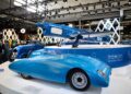 A blue streamlined car and a blue vintage aircraft, evoking European elegance, are displayed on a white platform at an indoor exhibition, with people and other vehicles visible in the background—an ambience reminiscent of Rétromobile NY in Manhattan.