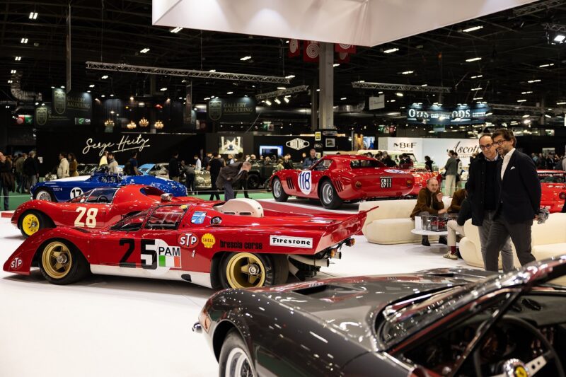 A group of vintage race cars is displayed indoors at Rétromobile NY, where several people walk around, soaking in the European elegance and rich automotive history on display.