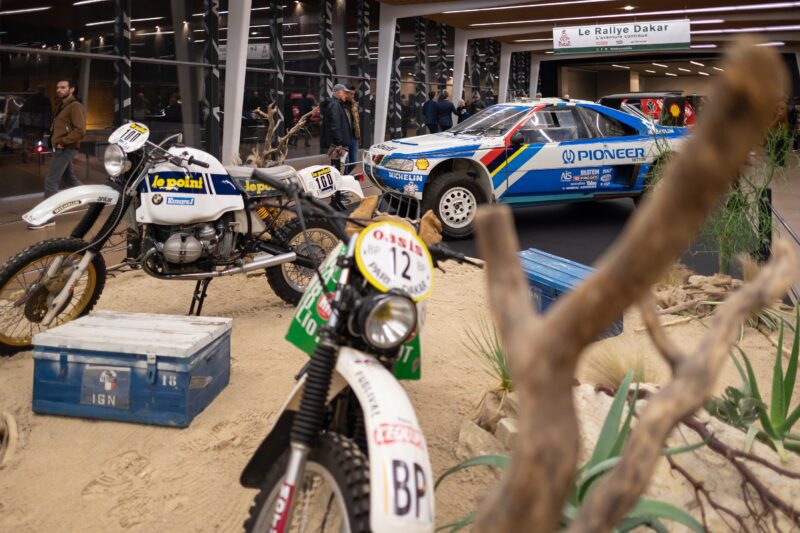 A museum exhibit in Manhattan showcases vintage rally vehicles, including a motorcycle and a Peugeot race car, blending European elegance with desert-themed decor beneath a "Le Rallye Dakar" sign—a nod to Rétromobile NY style.