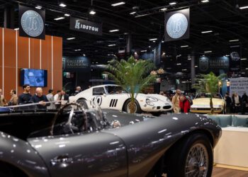 Experience European elegance at Rétromobile NY, an indoor Manhattan car show where visitors admire classic cars, including a silver convertible in the foreground and a white sports car on display in the background.