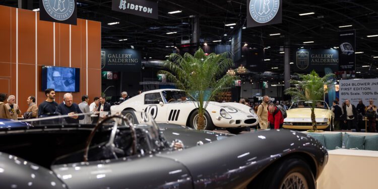 Experience European elegance at Rétromobile NY, an indoor Manhattan car show where visitors admire classic cars, including a silver convertible in the foreground and a white sports car on display in the background.