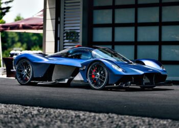 A Satin Mariana Blue Aston Martin Valkyrie hypercar is parked on a paved surface in front of a modern building.