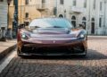 A maroon Pininfarina Battista sports car is parked on a cobblestone street in front of historic European-style buildings.