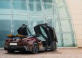 A burgundy Pininfarina Battista with scissor doors open is parked on a cobblestone surface in front of a modern glass building.