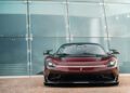 A maroon Pininfarina Battista with a black roof is parked in front of a modern glass building, photographed from the front.