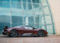 A maroon Pininfarina Battista with black accents is parked on a brick surface in front of a modern glass and stone building.