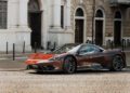 A sleek red Pininfarina Battista is parked on a cobblestone street in front of a historic building with arched windows and ornate details.