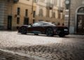 A dark red Pininfarina Battista sports car is parked on a cobblestone street in front of historic beige buildings with balconies and shuttered windows.