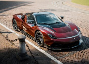A sleek red and black Pininfarina Battista sports car is parked on a cobblestone street near a curb lined with short metal posts and chains.