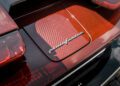 Close-up of a red carbon fiber Pininfarina Battista car with a herringbone pattern and a silver "Pininfarina" badge on the rear.