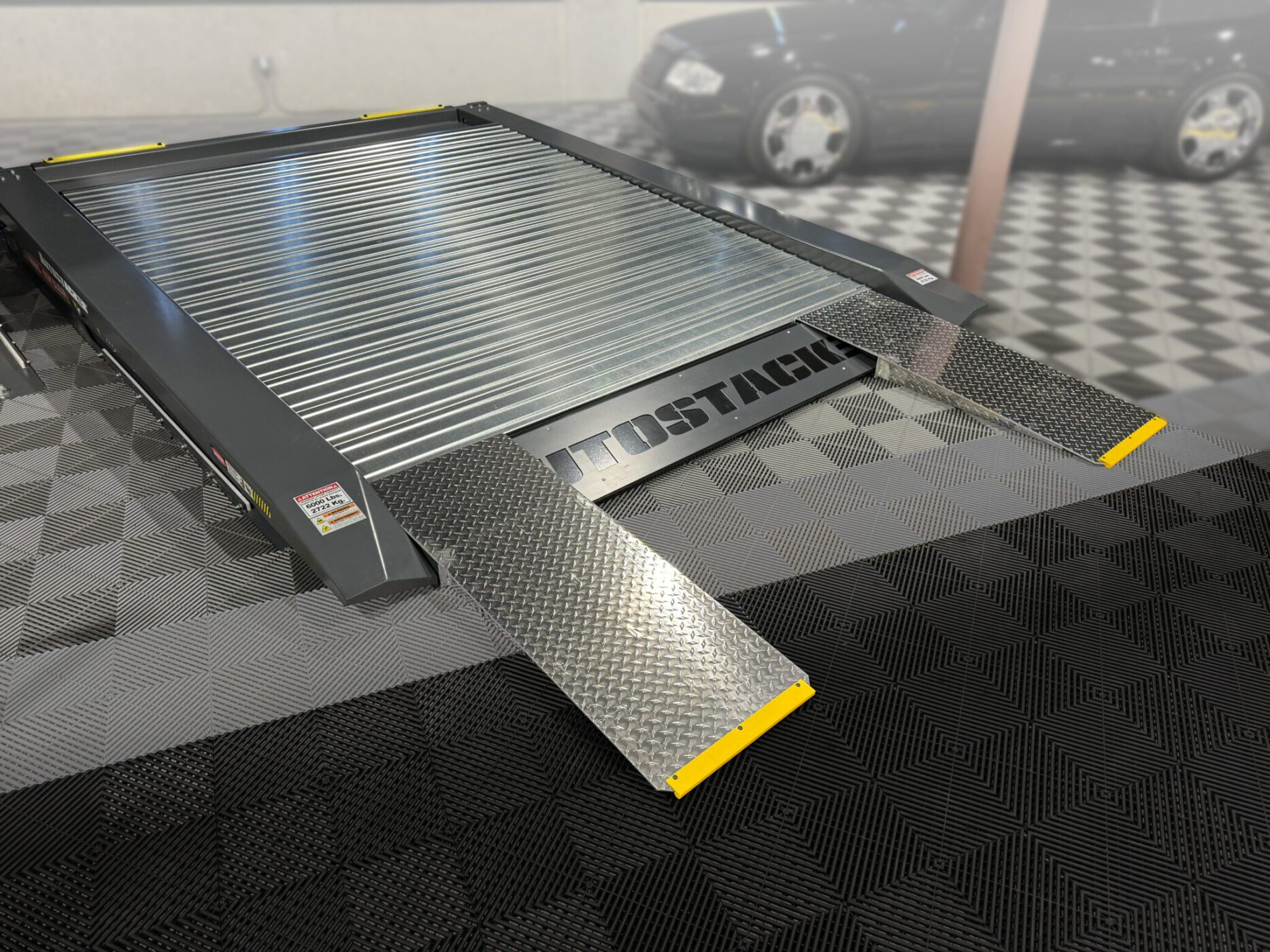 Metal parking platform with two diamond-plate lift ramps and "AUTOSTACKER" branding, positioned on a checkered floor in a garage with an Autostacker Lift and a blurred car in the background.