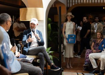 A man in a cap speaks into a microphone during an indoor event at the Miami Concours 2025, with an audience seated around him and a woman standing nearby holding a blue box, highlighting the essence of automotive luxury.