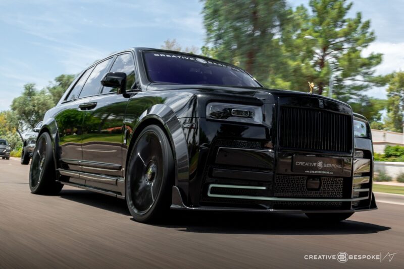 A black luxury SUV with tinted windows and custom detailing, a true unique garage creation, drives on a sunny, tree-lined street.