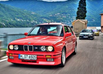 A red BMW E30 M3 drives along a lakeside road with mountains in the background, followed by another important car.