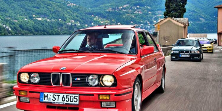 A red BMW E30 M3 drives along a lakeside road with mountains in the background, followed by another important car.