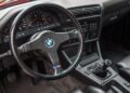 BMW car interior featuring a black leather steering wheel, manual gear shift, analog gauges, and dashboard controls—hallmarks of important BMWs like the legendary E30 M3.