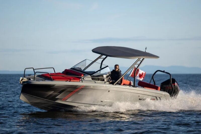 A person drives a gray motorboat with a canopy and red seating on open water, showcasing a true maritime experience, with a Canadian flag visible at the rear.