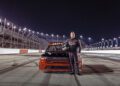 A race car driver in a racing suit stands holding a helmet in front of a red and black Ram truck powered by HEMI V8s on a racetrack at night.