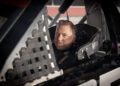 A man wearing a racing suit and helmet sits in the driver's seat of a NASCAR Ram, looking out through the window netting.