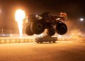 A monster truck jumps over a pickup truck on a racetrack at night, with large flames shooting up in the background—hinting at the excitement of Ram NASCAR return and roaring HEMI V8s in 2026.