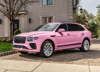 A Pink Bentayga SUV is parked on a brick driveway in front of a beige house with landscaped greenery, showing off one of the unique Bentley exterior colors.