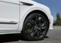 Close-up of a new Bentayga’s front wheel, featuring black alloy rims with a “B” logo, green brake calipers, and a "Speed" badge on the fender. The powerful Bentayga stands out against trees and sky in the background.