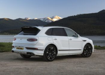 A powerful Bentayga, this white luxury SUV is parked on a dirt road by a lake, with mountains and trees in the background under a clear sky.
