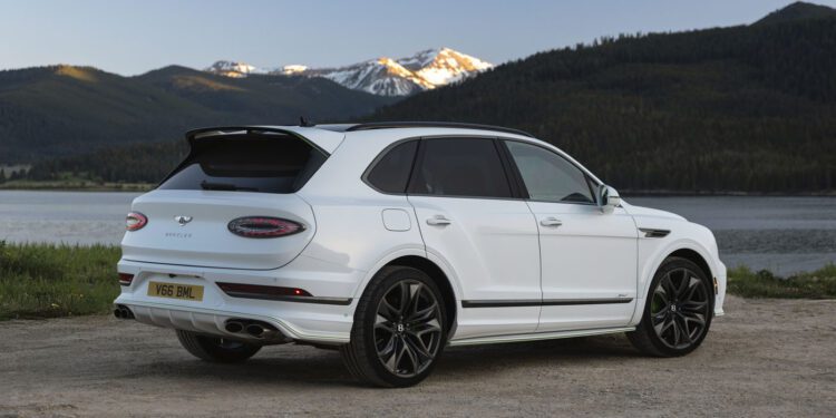 A powerful Bentayga, this white luxury SUV is parked on a dirt road by a lake, with mountains and trees in the background under a clear sky.