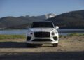 A white Bentley Bentayga Speed luxury SUV is parked on a dirt surface near a lake, with forested hills and snow-capped mountains in the background.