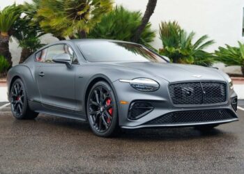 A sleek matte gray luxury sports coupe in stealth mode is parked on wet pavement, framed by palm trees and lush greenery.