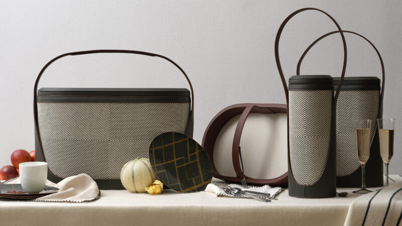 A set from the Hyde Picnic Collection features woven baskets, a plate, cutlery, two champagne flutes, apples, a small pumpkin, and a cup arranged on a cream tablecloth—perfect for refined outdoor living by Bentley Home.