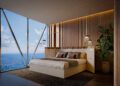 Modern penthouse bedroom at Bentley Residences Miami with a large bed, wooden accents, pendant lights, a potted plant, and floor-to-ceiling windows overlooking the ocean and sky.