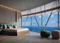 A modern penthouse bedroom with a large bed, two ottomans, and floor-to-ceiling windows offering an ocean view under a partly cloudy Miami sky.