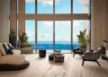 Modern penthouse living room with large floor-to-ceiling windows, beige sofas, elegant chairs, potted plants, and a stunning ocean view under a blue sky—an exquisite example of Miami real estate at Bentley Residences Miami.