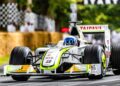 A Formula 1 race car with prominent sponsorship logos speeds down a track lined with hay bales and checkered flags, celebrating 75 Years Of F1 at the Festival of Speed.