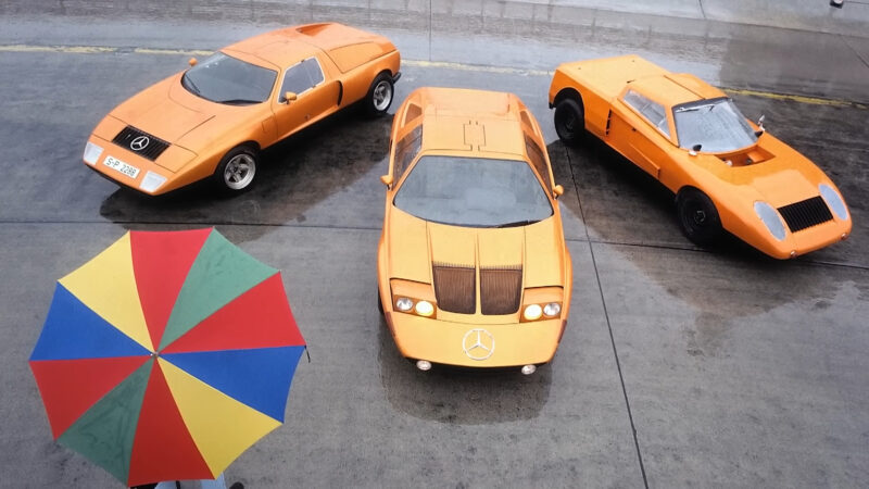 Three orange vintage Mercedes-Benz sports cars, including a striking Vision One-Eleven concept, parked on wet pavement and viewed from above, with a colorful umbrella in the foreground.