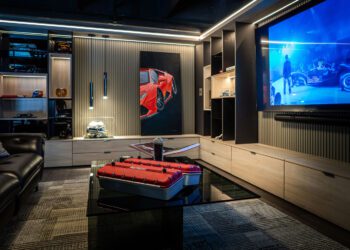 Modern entertainment room with a large wall-mounted TV showing a racecar, a black leather sofa, car-themed decor, and a glass coffee table displaying automotive items—perfect for Palm Beach luxury car collectors or CollectionSuites enthusiasts.
