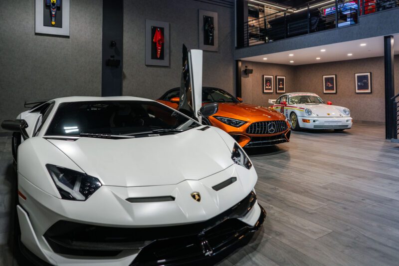 Three luxury sports cars—a white Lamborghini, an orange Mercedes-Benz, and a white Porsche—are parked inside a modern, well-lit CollectionSuites garage with framed automotive art, perfect for Palm Beach luxury car collectors.