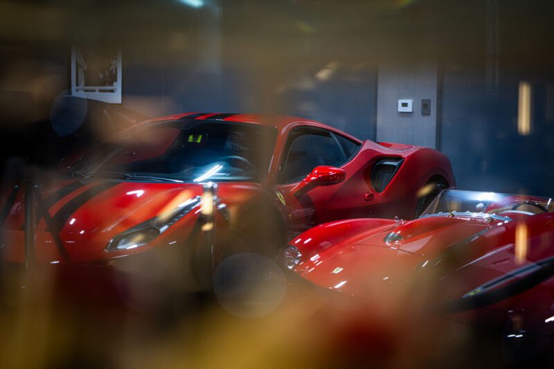 Two red sports cars, including a Ferrari, are parked indoors at CollectionSuites Palm Beach, reflecting the allure sought by luxury car collectors amid blurred lights in the foreground.