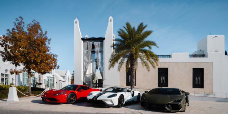 Three luxury sports cars—a red, a white, and a dark green—are parked side by side in front of a modern white building at Alys Beach, capturing The Art of the Drive beneath swaying palms and a clear blue sky.