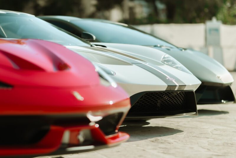 Three sports cars, including a red and two silver models, are parked side by side in a sunlit outdoor setting at the Art of the Drive, part of Cars of 30A in Alys Beach.