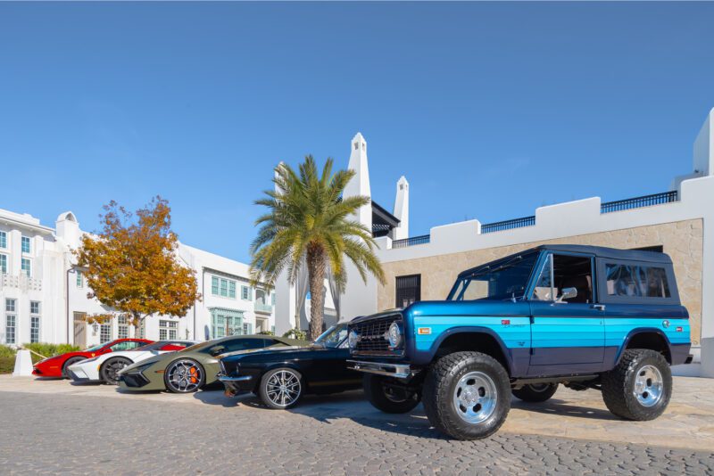 A blue vintage Ford Bronco is parked beside three modern sports cars at a car show in Alys Beach, with palm trees and white buildings under a clear blue sky.