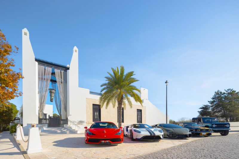 Four luxury cars, including a red Ferrari and a classic blue convertible, are parked in front of a modern white building with a palm tree at Alys Beach—capturing the essence of Cars of 30A and the Art of the Drive.