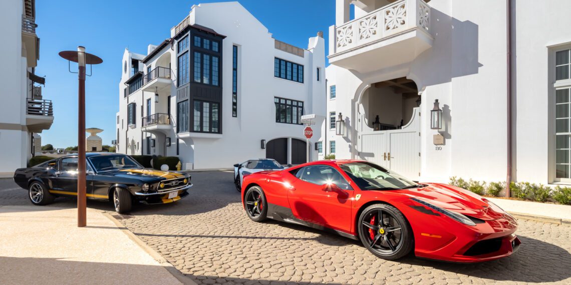 A red Ferrari and a black classic car parked on a cobblestone street in Alys Beach showcase the Art of the Drive, set against modern white buildings beneath a clear blue sky—signature moments from Cars of 30A.