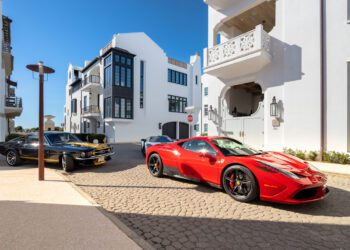 A red Ferrari and a black classic car parked on a cobblestone street in Alys Beach showcase the Art of the Drive, set against modern white buildings beneath a clear blue sky—signature moments from Cars of 30A.