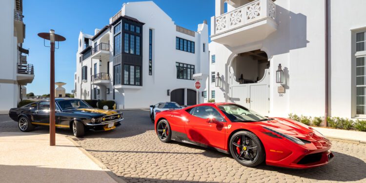 A red Ferrari and a black classic car parked on a cobblestone street in Alys Beach showcase the Art of the Drive, set against modern white buildings beneath a clear blue sky—signature moments from Cars of 30A.