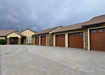 Large single-story house with a tan exterior, stone accents, and four wooden garage doors sits on a wide concrete driveway under a cloudy sky—offering unique opportunities through CWS Marketing Group.