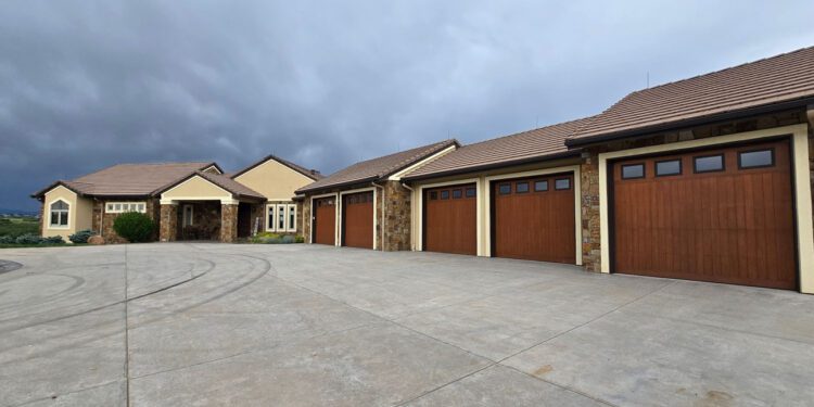 Large single-story house with a tan exterior, stone accents, and four wooden garage doors sits on a wide concrete driveway under a cloudy sky—offering unique opportunities through CWS Marketing Group.