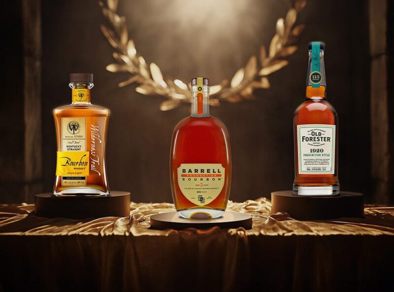Three bourbon whiskey bottles—Wilderness Trail, Barrell Bourbon, and Old Forester 1920—are displayed on pedestals against a dark background with a golden laurel wreath, perfect picks for the duPont REGISTRY Father's Day Gift Guide.